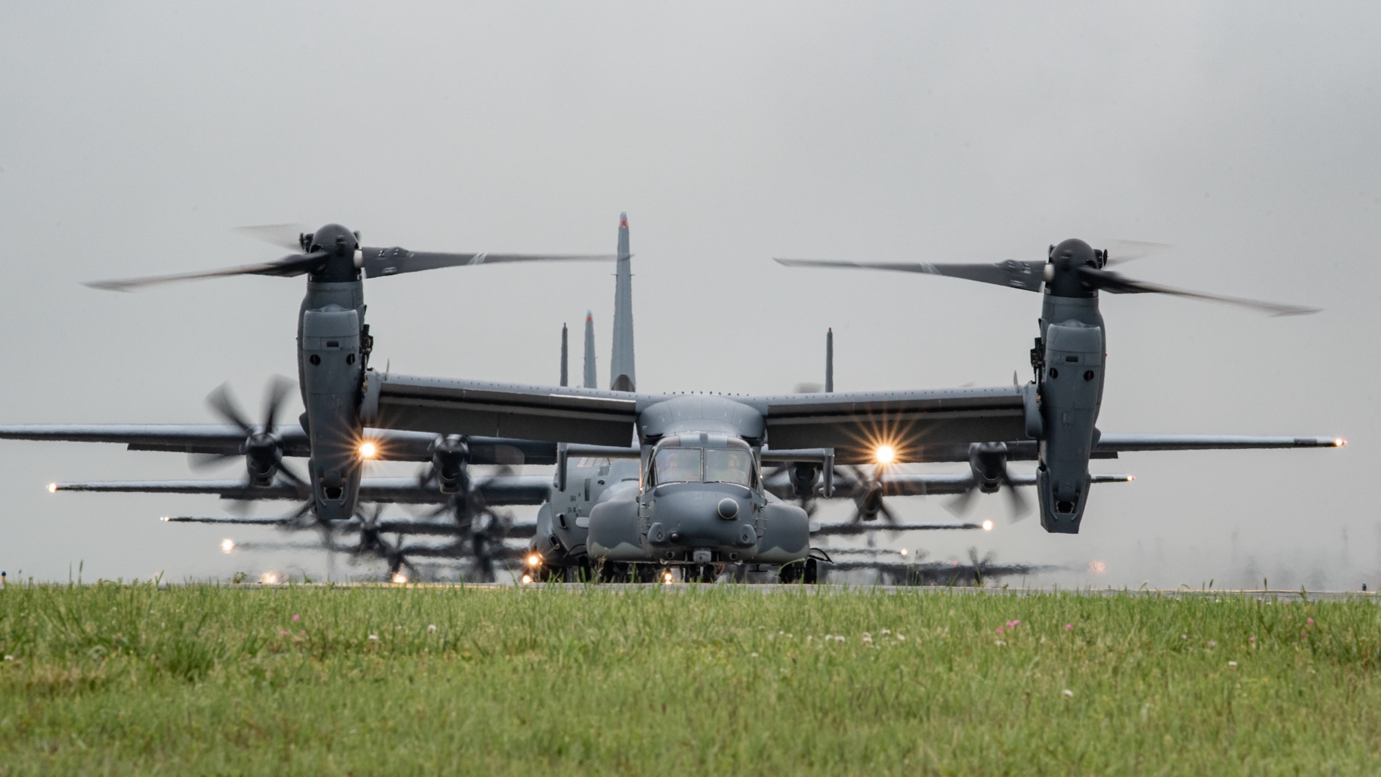A US Air Force CV-22 Osprey taxiing with its nacelles vertical and rotors turning, with MC-130 aircraft lined up behind it